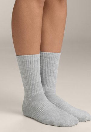 Par svijetlosivih rebrastih čarapa do sredine lista koje se nose na golim nogama, stoje na neutralnoj površini s jednostavnom pozadinom.