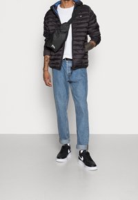 Osoba ubrana w czarną kurtkę puchową na białej koszuli, jasnoniebieskie podwinięte dżinsy, czarno-białe trampki oraz niosąca czarną torbę na ramię.