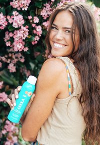 Turkusowa butelka sprayu przeciwsłonecznego z białą nakrętką, oznaczona "COOLA", trzymana na tle różowych kwiatów, z żebrowanym beżowym wierzchem.