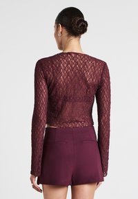 Top corto in pizzo borgogna con maniche lunghe, con un design trasparente, abbinato a pantaloni corti a vita alta coordinate. Motivo floreale testurizzato visibile.