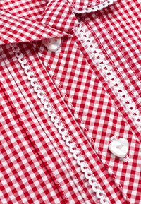 Camicia a quadretti rosso e bianco con chiusura frontale con bottoni, passamaneria bianca decorativa e bottoni a forma di cuore. Tessuto in cotone, colletto classico.