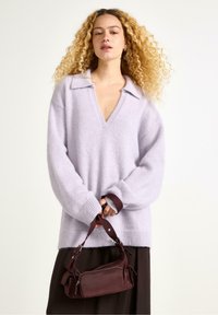 Donna con capelli biondi ricci indossa un maglione leggero viola a V e tiene in mano una piccola borsa bordeaux su uno sfondo semplice.