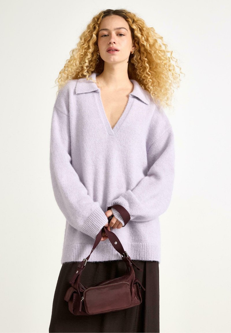 Donna con capelli biondi ricci indossa un maglione leggero viola a V e tiene in mano una piccola borsa bordeaux su uno sfondo semplice.