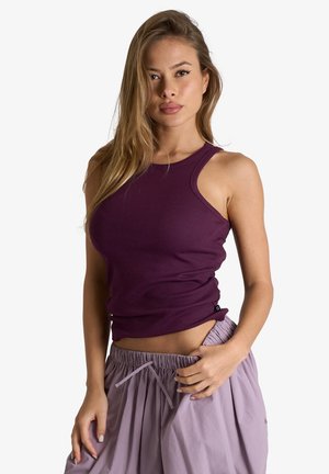 Donna con i capelli lunghi che indossa un top viola senza maniche a coste e pantaloni viola chiaro con coulisse, posando su uno sfondo bianco semplice.