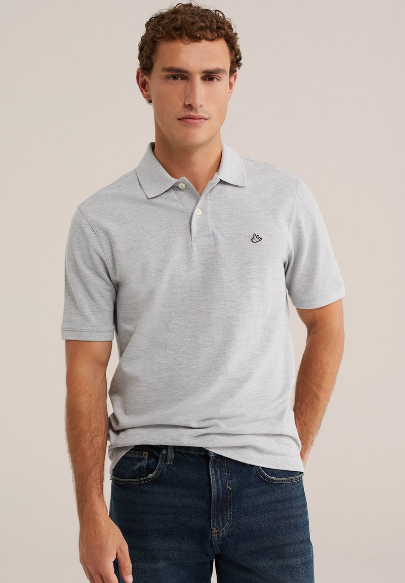 Jeune homme aux cheveux bouclés portant un polo gris clair à manches courtes et un jean bleu foncé, debout devant un fond uni.