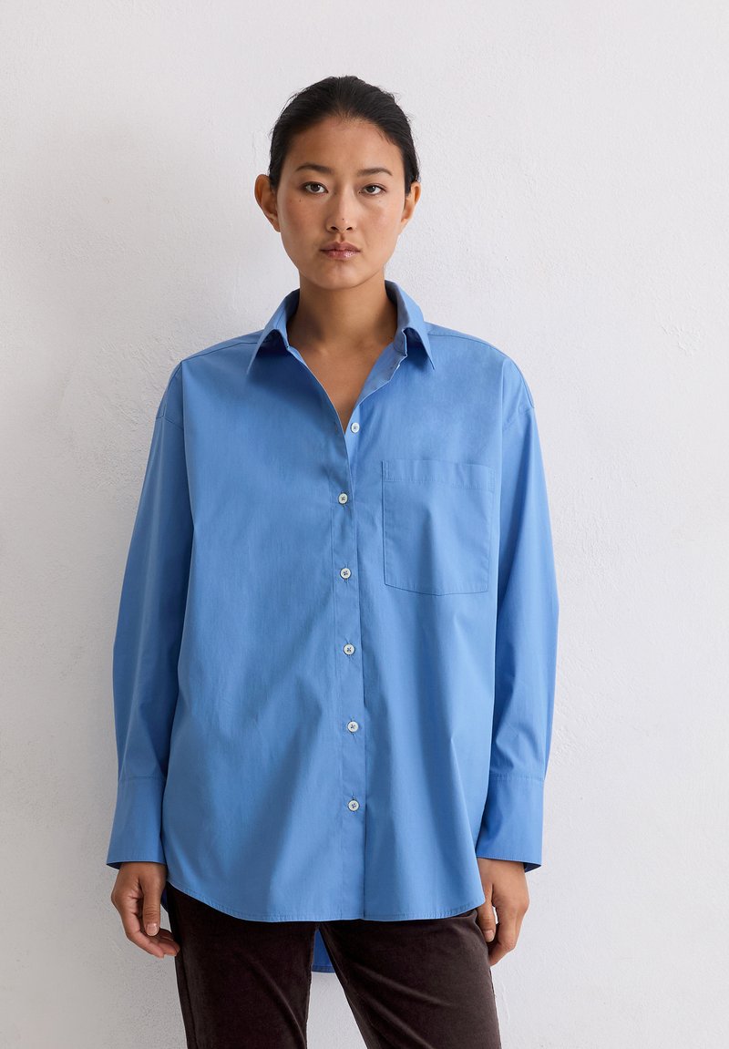 Vrouw staat tegen een witte muur, draagt een oversized lichtblauwe, knoopbare blouse en donkere broek, kijkt recht vooruit met een neutrale uitdrukking.