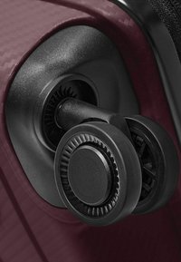 Primo piano delle ruote nere a doppio spinner di una valigia attaccate all'angolo di una valigia di colore rosso scuro con texture.
