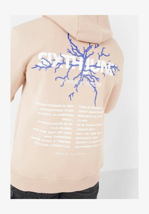 Perzikhuiden met een blauwe boomafbeelding en witte tekst. De achterkant heeft zinnen over streetwear en bevat de datum "6 JUNI 2008."