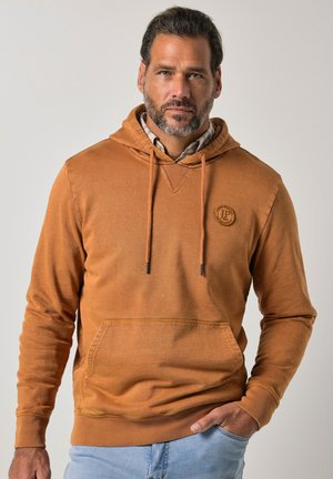 Man met baard die een lichtbruine hoodie en lichtblauwe spijkerbroek draagt, staand met één hand in zijn zak tegen een effen achtergrond.