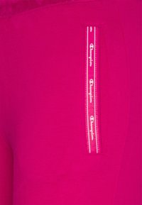 Felle roze stof met een ritszak en een wit "Champion" logo. De focus ligt op de details van de zak en de levendige kleur.