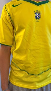 Żółta koszulka Nike z zielonymi akcentami, logo CBF i napisem Brasil. Posiada dolną część z siateczki i gładką teksturę. Krótkie rękawy.