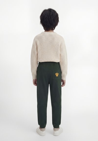 Παιδί με κοντά σκούρα μαλλιά στέκεται με πρόσωπο προς ένα λευκό φόντο, φορώντας μπεζ πλεκτό πουλόβερ, σκούρο πράσινο παντελόνι με χρυσό έμβλημα και λευκά αθλητικά παπούτσια.