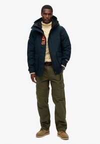 Giacca trapuntata blu navy con un grande logo arancione, maglione a maglia crema, pantaloni cargo verdi oliva e scarpe color crema. Presenta zip e tasche.