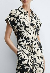 Vestido negro de manga corta con un patrón floral blanco, botonadura frontal, dos bolsillos en el pecho y cintura atada. Confeccionado con una tela ligera.