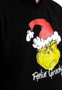 Felpa nera con cappuccio che presenta una grafica del Grinch con un cappello di Babbo Natale rosso con bordi bianchi, viso giallo acceso e testo "Feelin' Grinchy" in bianco.