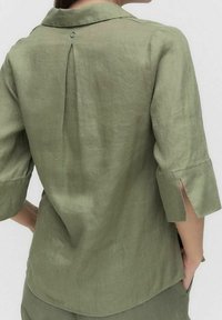 Vihreä pellavapaita, jossa on kaulus, 3/4-hihate, takana laskos ja pyöreä metallinappi yksityiskohtana kaulassa.