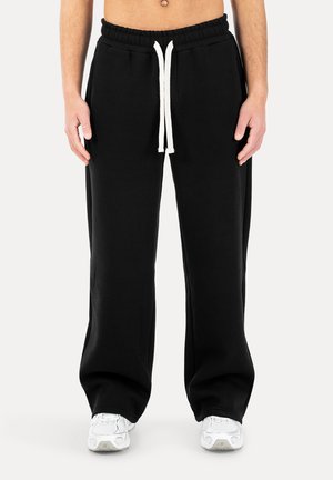 Schwarze, locker sitzende Jogginghose mit weißem Kordelzug, getragen mit weißen Sneakers an einer stehenden Person vor einem schlichten Hintergrund.