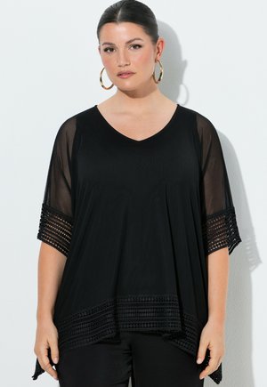 Femme aux cheveux tirés en arrière, portant de grandes boucles d'oreilles en or, un haut noir ample avec des manches transparentes et une bordure à motifs, posant devant un mur blanc.