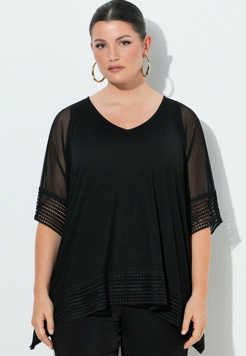 Kobieta z włosami zaczesanymi do tyłu, nosząca duże złote kolczyki-koła oraz luźną czarną bluzkę z przezroczystymi rękawami i wzorzystymi wykończeniami, stoi przy białej ścianie.