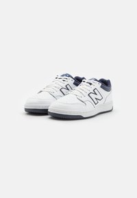 Białe skórzane sneakersy z granatowymi akcentami, detale z perforacji, okrągłe sznurowadła i gumowe podeszwy. Posiadają wyraźne logo "N" po bokach.