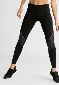 Žena stojící se široce rozkročenýma nohama, oblečená v černých legínách s tmavě šedými panely a černými sportovními botami na světlém pozadí.