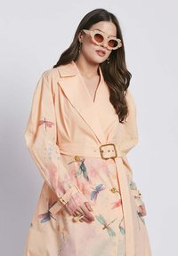 Trench coat color pesca con vita a vita regolabile; presenta motivi floreali di libellule e accenti ad acquerello. Maniche lunghe con dettagli a bottone.