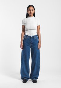 Camiseta blanca de manga corta con diseño de cuello alto, combinada con jeans de mezclilla azul de corte holgado. Zapatos negros completan el conjunto.