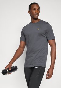 Pilkas medvilnės marškinėliai su apvalia iškirpte ir trumpomis rankovėmis, su mažuoju aukso logotipu ant krūtinės. Modelis laiko juodą vandens butelį.