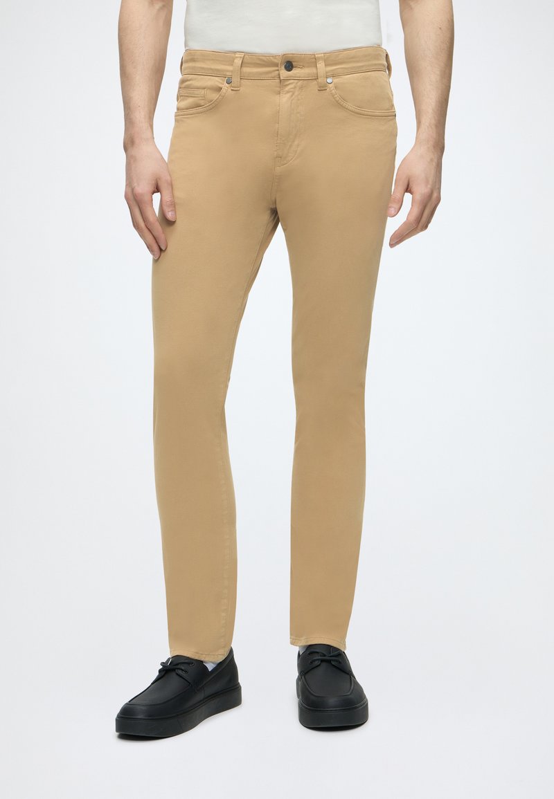 Homme portant un pantalon beige slim avec poches avant et des chaussures noires à lacets, debout devant un fond blanc uni.
