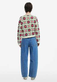 Virkat tröja med blommiga rutor i rosa och grönt på en krämfärgad bakgrund; lös passform, tillsammans med vida blå jeans.
