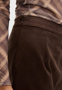 Hnědé semišové kalhoty s hladkou texturou a přiléhavým střihem. Obsahují diskrétní boční zip a čistý design pasu.