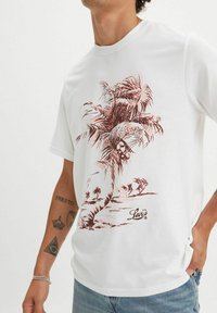 Camiseta de algodón blanca con un gráfico de palmera roja y el logo de "Levi's" en la parte inferior delantera. Manga corta y escote redondo.