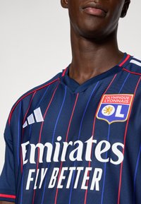 Tamsiai mėlyna futbolo marškinėliai su vertikaliomis raudonomis ir mėlynomis juostelėmis, kuriuose yra balti tekstai "Emirates Fly Better" ir siuvinėtas komandos logotipas.
