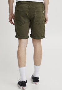 Pantaloni scurți din denim verde măsliniu cu tiv răsucit, având două buzunare la spate și închidere cu fermoar, asortați cu șosete albe și adidași negri.