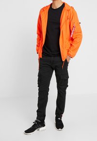 Ljust orange lättviktsjacka med huva, dragkedja och fickdetaljer, ihop med svarta cargobyxor och svarta sportskor.