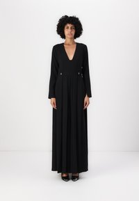 Schwarzes langärmliges Kleid mit tiefem V-Ausschnitt und plissiertem Rock. Verfügt über zwei runde Akzente an der Taille. Strukturierter Stoff und volle Länge.