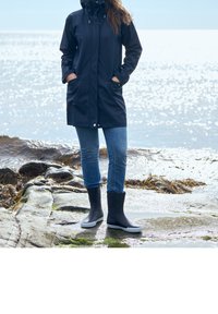 Imperméable bleu marine avec capuche et deux poches avant, associé à un jean bleu et des bottes en caoutchouc noires avec semelles blanches, sur une côte rocheuse.