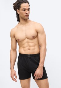 Homme musclé avec des dreadlocks portant un short noir, regardant vers la gauche, debout devant un fond clair uni.