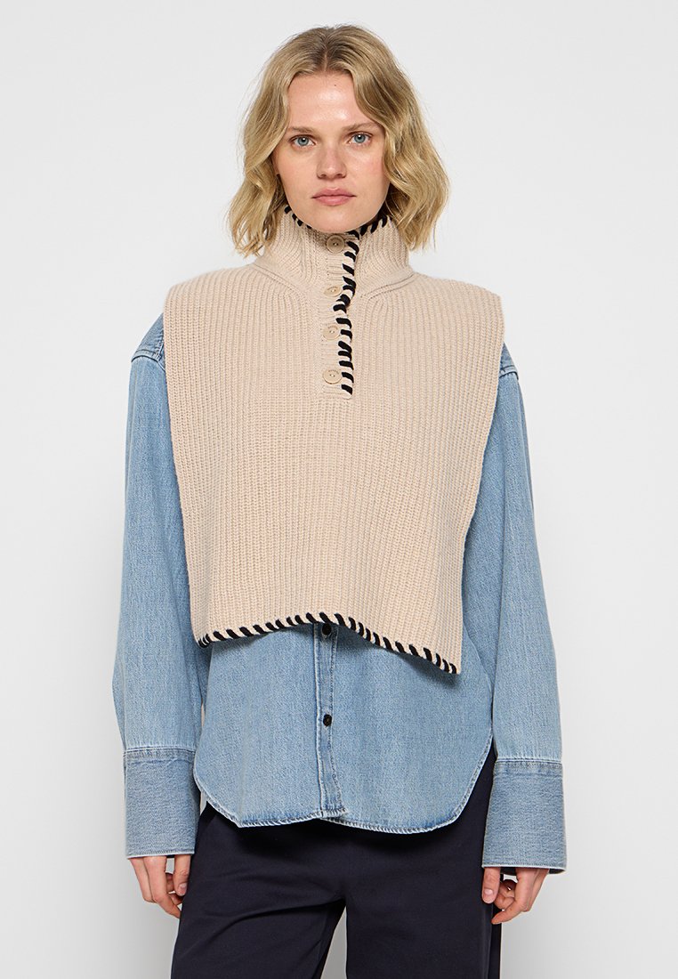 Beige gestrickte ärmellose Weste mit hohem Kragen, mit einer Knopfleiste und schwarzen gestreiften Akzenten an den Rändern, getragen über einem Jeanshemd.