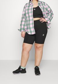 Kvinna klädd i svart Adidas crop top och cykelshorts med svarta sneakers, mintgröna strumpor och en grön- och rosa-rutig skjorta.