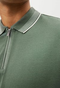 Vihreä tekstuurinen polo-paita, jossa on vetoketjukaulus. Kauluksen reunassa on valkoisen ja vihreän värin raita-akseli.