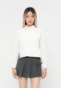 Hvit crop top med øyeletdesign og bølget kanter, med knappelukking og lange, luftige ermer. Kombinert med et grått, plissert skjørt.