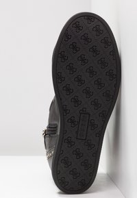 Suela de zapato de cuero negro con un patrón texturizado que presenta logotipos. Cremalleras laterales y detalles con tachuelas visibles a lo largo del borde lateral. Base plana y sólida.