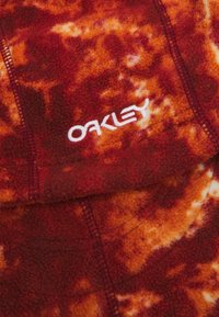 Tela de forro de polar con un patrón en rojo y naranja que presenta un bordado blanco con el logo "Oakley" y costuras oscuras contrastantes.