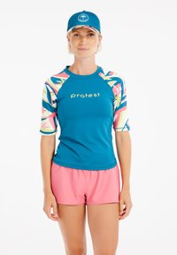 Teal rash guard dengan lengan bermotif berwarna-warni dan logo emas, dipadukan dengan celana pendek pink dan topi biru yang serasi dengan logo.
