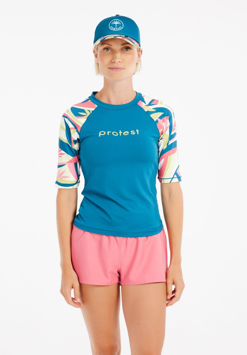 Teal rash guard dengan lengan bermotif berwarna-warni dan logo emas, dipadukan dengan celana pendek pink dan topi biru yang serasi dengan logo.