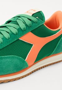 Grønn sneaker med mesh- og semsket skinnmaterialer, med en lys orange aksent og klassisk snøring. Gummisåle med strukturert grep.