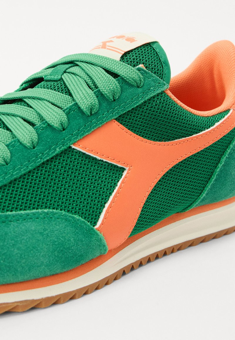 Grüner Sneaker aus Mesh- und Wildledermaterialien, mit einem leuchtend orangefarbenen Akzent und klassischem Schnürdesign. Gummi-Sohle mit strukturiertem Griff.