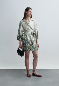 Robe-chemise à motif floral en crème clair, dotée d'une ceinture à la taille et de manches larges ; accompagnée d'un sac à main noir et de ballerines bordeaux.