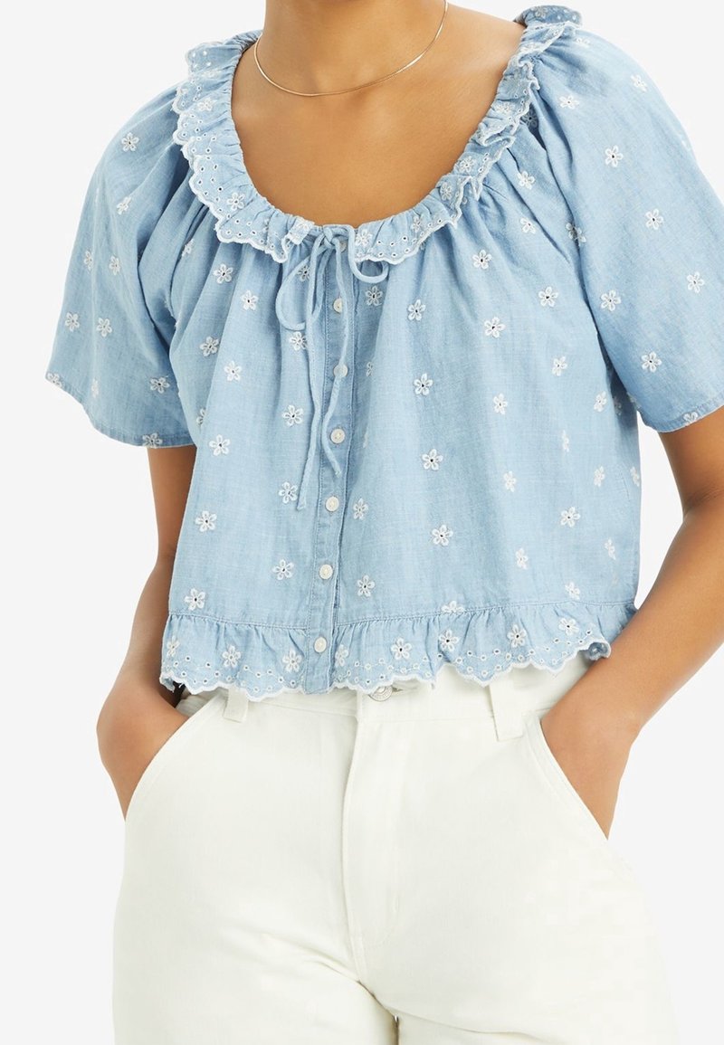 Blouse courte à manches courtes bleu clair avec un col froncé, broderies florales, boutons sur le devant et dentelle au niveau de l'ourlet. Portée avec un pantalon blanc.